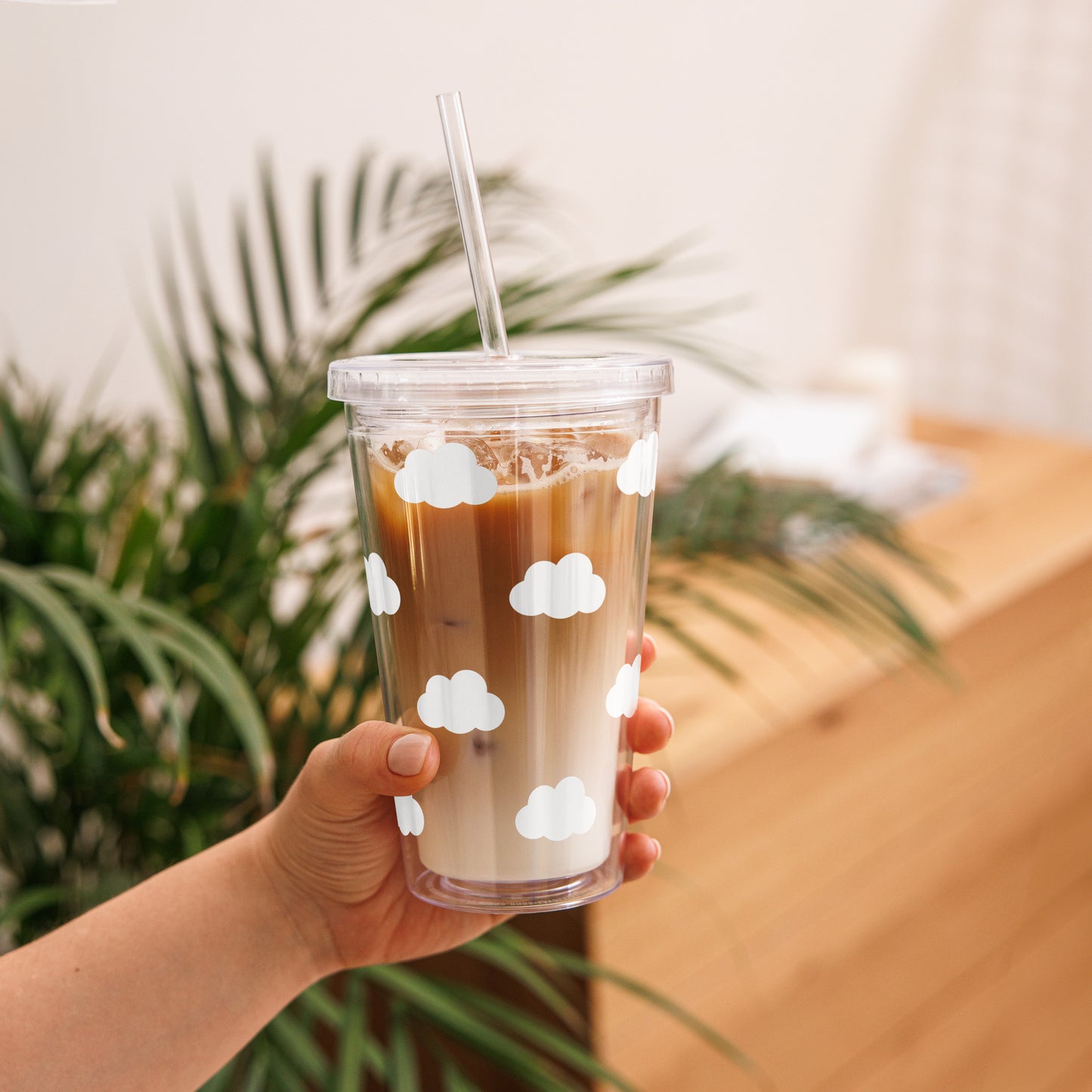 Hand holding a clear cup with iced coffee and cloud designs, against a blurred indoor background.