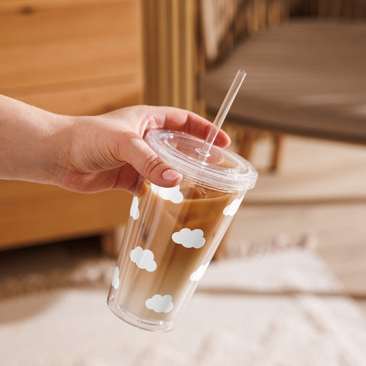 Hand holding a clear cup with a straw, filled with a creamy beverage, against a blurred indoor background.