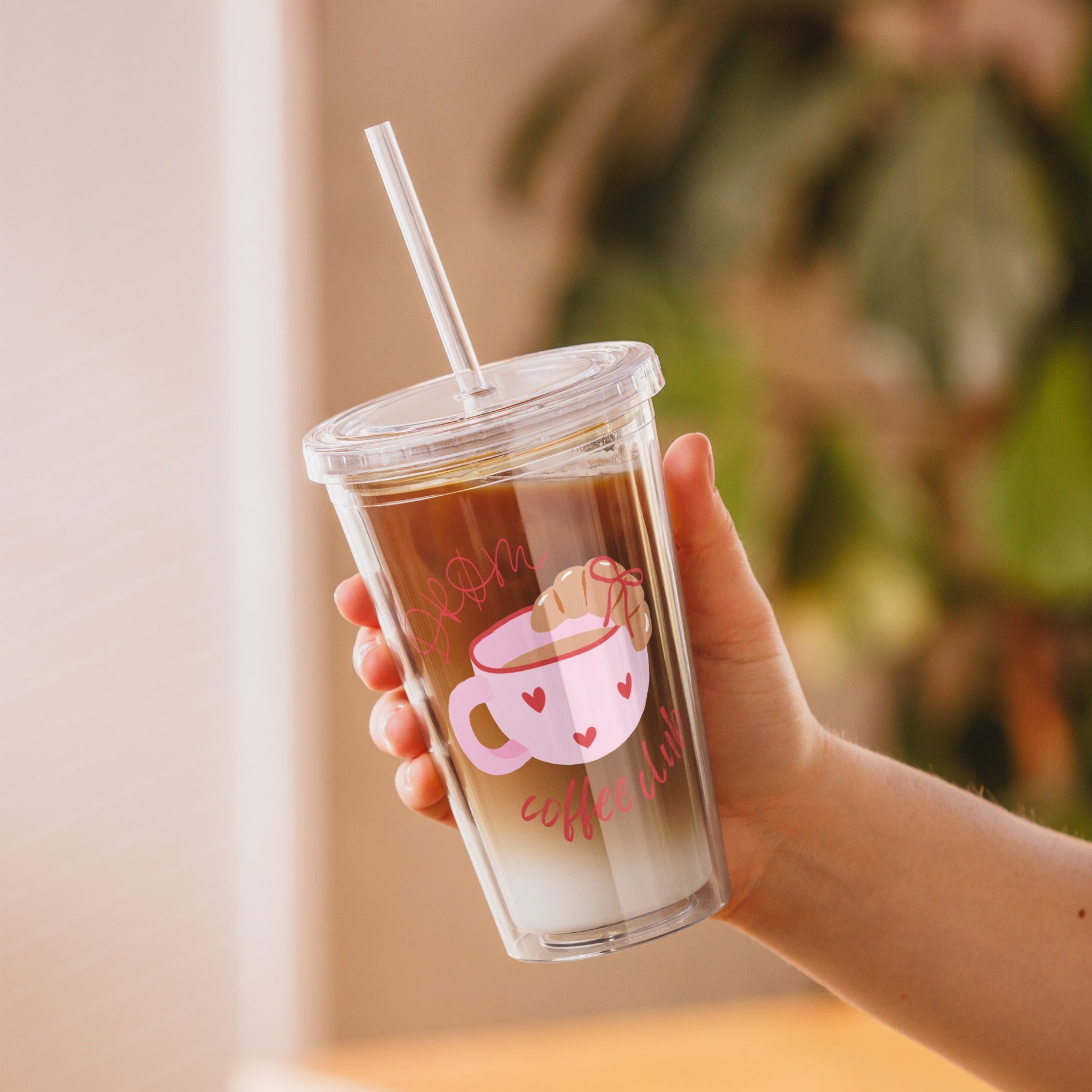 Hand holding a clear tumbler with a design of a pink coffee cup and text, filled with a beverage.