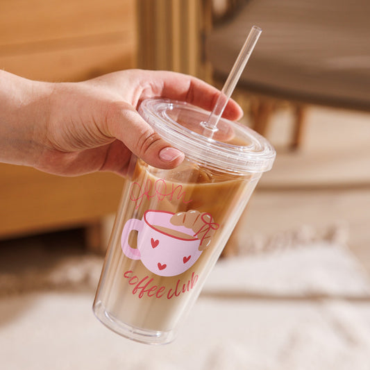 Hand holding a clear cup with a straw, featuring a pink coffee cup design, against a blurred indoor background.