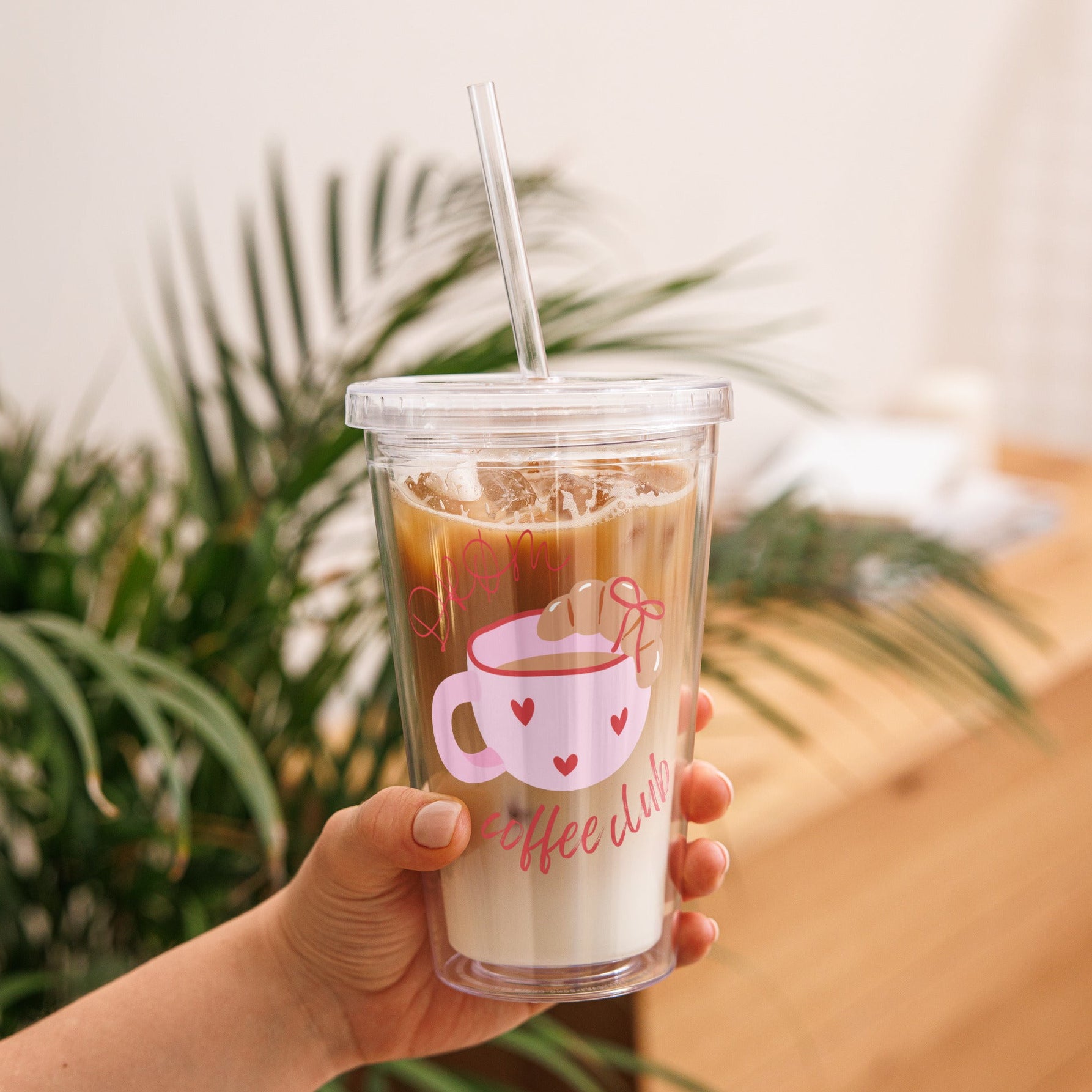 Hand holding a clear cup with iced coffee and a straw, against a blurred indoor background with plants.