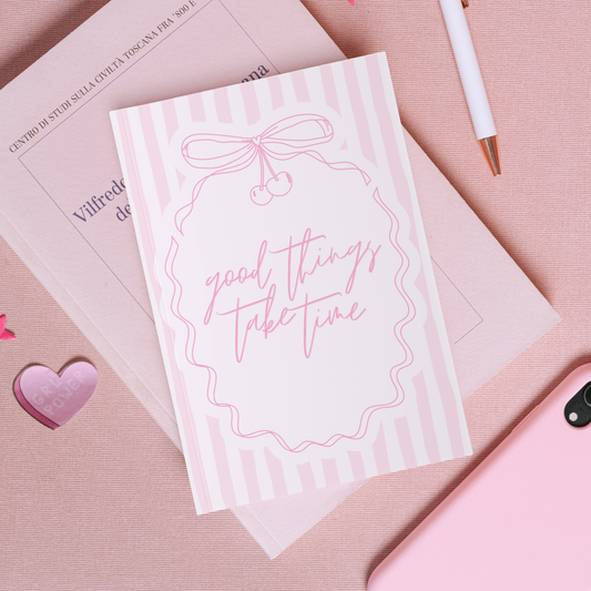 Notebook with 'good things take time' text on a pink surface with a heart and pen.