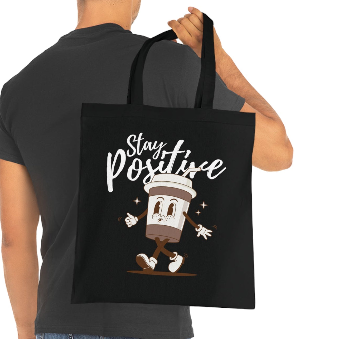 Black tote bag with a cartoon coffee cup and 'Stay Positive' text, held by a person wearing a black t-shirt.